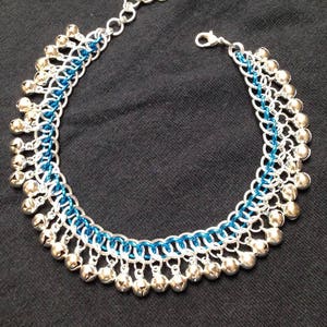 May include: A silver chain anklet with blue accents and silver bells. The anklet is made of metal and has a lobster clasp closure.
