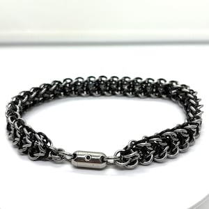 May include: A silver and black chainmail bracelet. The bracelet is made of interlocking metal rings, creating a textured surface. It features a silver clasp closure. The jewelry is displayed on a white surface.