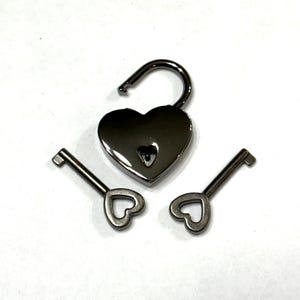 May include: A heart-shaped padlock with an open shackle and two matching keys. The lock and keys are silver-colored and have heart-shaped details. The lock is open, suggesting access or security.