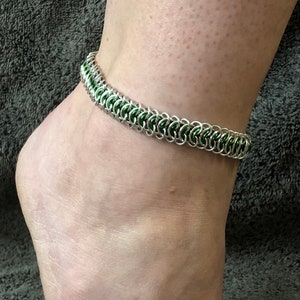 May include: A silver and green chainmail ankle bracelet. The bracelet is made of small, interlocking metal rings.