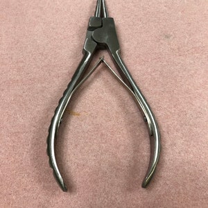 May include: A pair of silver-colored round nose pliers. The pliers have a ribbed handle for grip and are designed for jewelry making or crafting. The tool is shown against a pink background.