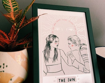May include: A framed print with a light pink background featuring a black and white illustration of two people. The text "Of the stars, and dust, that is the sun" is written in a cursive font above the illustration. The text "THE SUN" is written in a bold font below the illustration.