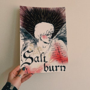 May include: An art print featuring a stylized illustration of a person with angel wings, blush, and a halo. The print has the word "Saltburn" in gothic lettering, with handwritten text and floral accents. The color palette includes gold, red, and black.