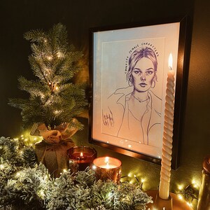 Op de afbeelding: Een zwart-wit portret in een lijst van een vrouw met de tekst "Complex Female Characters". Een kleine kerstboom met lichtjes, twee brandende kaarsen en een slinger van groen met lichtjes staan op de voorgrond.
