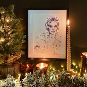Op de afbeelding: Een ingelijste potloodschets van een vrouw met de tekst "COMPLEX FEMALE CHARACTERS" wordt op een schoorsteenmantel getoond. Een brandende kaars en een kleine kerstboom staan op de schoorsteenmantel. De sc&egrave;ne wordt warm verlicht door kaarsen en fee&euml;rieke lichtjes.