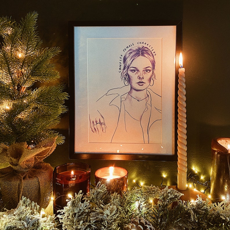 Op de afbeelding: Een ingelijste potloodschets van een vrouw met de tekst "COMPLEX FEMALE CHARACTERS" wordt op een schoorsteenmantel getoond. Een brandende kaars en een kleine kerstboom staan op de schoorsteenmantel. De sc&egrave;ne wordt warm verlicht door kaarsen en fee&euml;rieke lichtjes.
