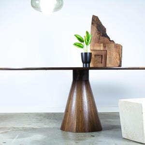 May include: A large, oval, dark wood dining table with a unique, conical base. The table is set on a concrete floor with a white, square stool in the foreground.