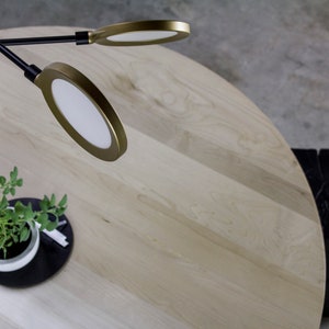 May include: A modern interior scene featuring a light wood round table, a small potted plant, and a black geometric side table. Overhead, a black and gold-toned light fixture with circular lights illuminates the scene.