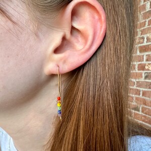 May include: A gold earring with a rainbow of small beads hanging from it. The earring is worn in the left ear of a person with long brown hair.