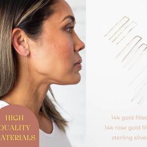 May include: A close-up of a person wearing long, thin, gold earrings. The earrings are made of 14k gold filled, 14k rose gold filled, and sterling silver. The text "HIGH QUALITY MATERIALS" is visible in the image.
