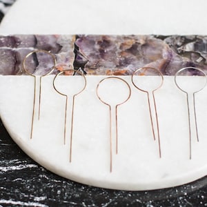 May include: Six gold hair pins with circular tops on a white marble surface. The surface has a purple and white agate inlay.