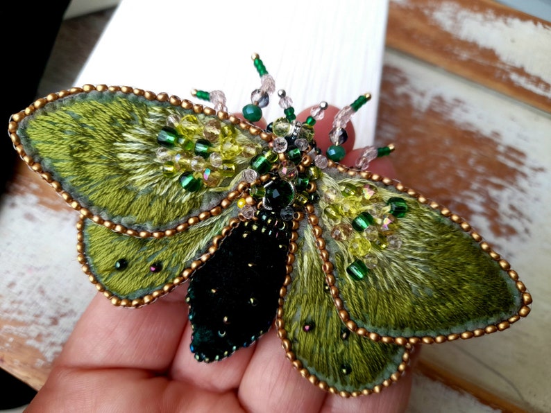 May include: A handcrafted, embroidered insect brooch. The wings are a textured green with gold bead edging. The body is black with a central black gem. The antennae are adorned with green and clear beads. A unique, wearable art piece.