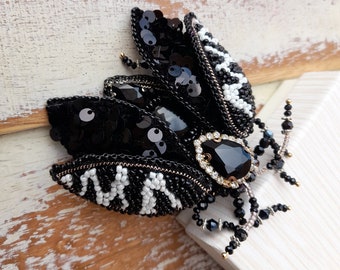 May include: A handcrafted insect-shaped brooch featuring black sequins, white seed beads, and a central faceted black teardrop stone surrounded by clear crystals. The wings display a textured pattern, and the legs are made of black and clear beads.