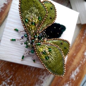 May include: A handcrafted, embroidered butterfly brooch. The wings are a vibrant green with gold bead edging and accents. The body features a dark green velvet center and is embellished with green and clear beads. The brooch is set on a white surface.