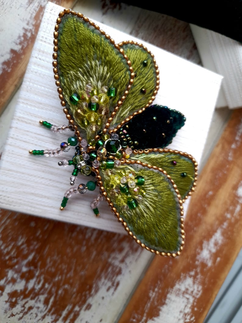 May include: A handcrafted, embroidered butterfly brooch. The wings are a vibrant green with gold bead edging and accents. The body features a dark green velvet center and is embellished with green and clear beads. The brooch is set on a white surface.