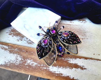 May include: A black and gold beaded insect brooch with purple and pink iridescent accents. The brooch is on a white marble surface.
