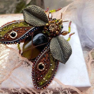 May include: A green and brown jeweled insect brooch with a large agate stone in the center. The wings are made of fabric and beads, and the body is made of metal.
