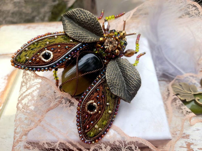 May include: A green and brown jeweled insect brooch with a large agate stone in the center. The wings are made of fabric and beads, and the body is made of metal.