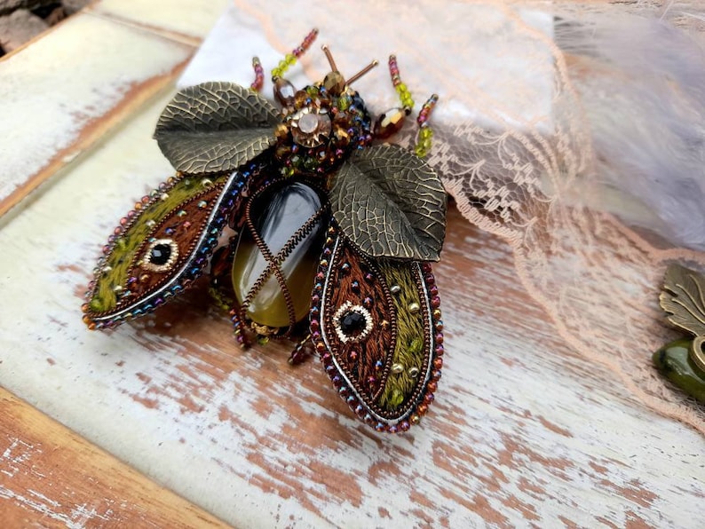 May include: A beaded and embroidered insect brooch with green, brown, and gold accents. The insect has a large, oval-shaped gemstone in the center.