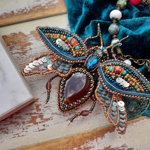 May include: A teal blue and copper colored beaded insect brooch with iridescent sequins and a large, clear, faceted gemstone in the center.