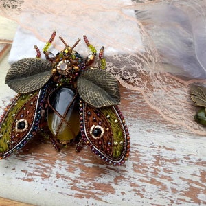 May include: A beaded and embroidered insect brooch with green, brown, and gold accents. The insect has a large, clear, faceted gemstone in the center of its body.