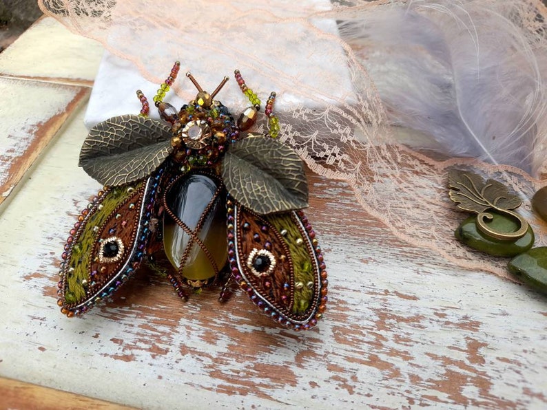 May include: A beaded and embroidered insect brooch with green, brown, and gold accents. The insect has a large, clear, faceted gemstone in the center of its body.