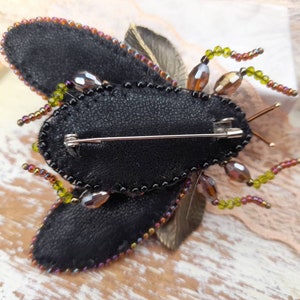 May include: A black and brown beaded insect brooch with a pin on the back. The insect has a black body with brown and green beads around the edges. The wings are made of black fabric and have a brown and green beaded trim.
