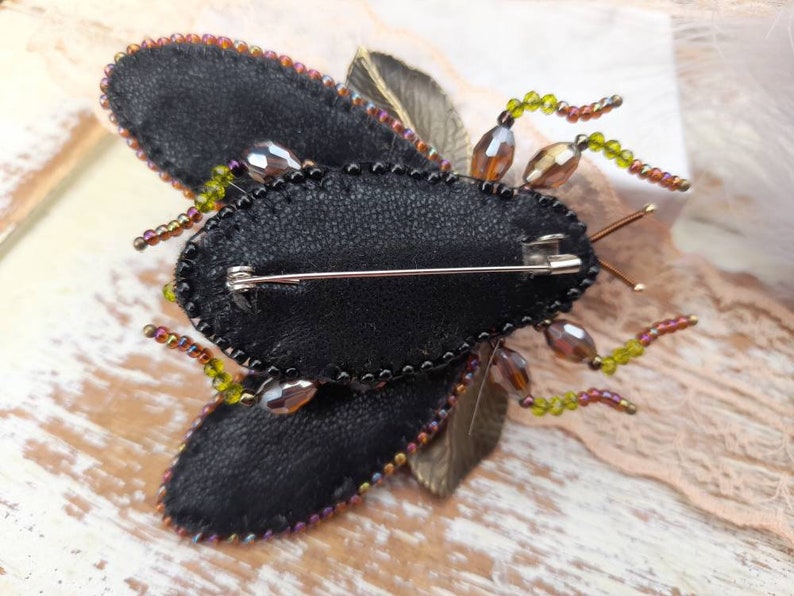 May include: A black and brown beaded insect brooch with a pin on the back. The insect has a black body with brown and green beads around the edges. The wings are made of black fabric and have a brown and green beaded trim.