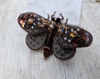 May include: A handcrafted insect-shaped brooch featuring a central brown banded agate stone body. The wings are embellished with dark grey velvet, metallic copper sequins, amber-colored crystals, and small white beads, finished with a beaded edge.
