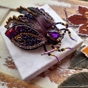 May include: A handcrafted beetle brooch featuring deep purple and black sequins, intricate beadwork, and a central faceted purple crystal. The insect body is accented with dark fur-like texture and delicate wire legs adorned with small purple and gold beads.