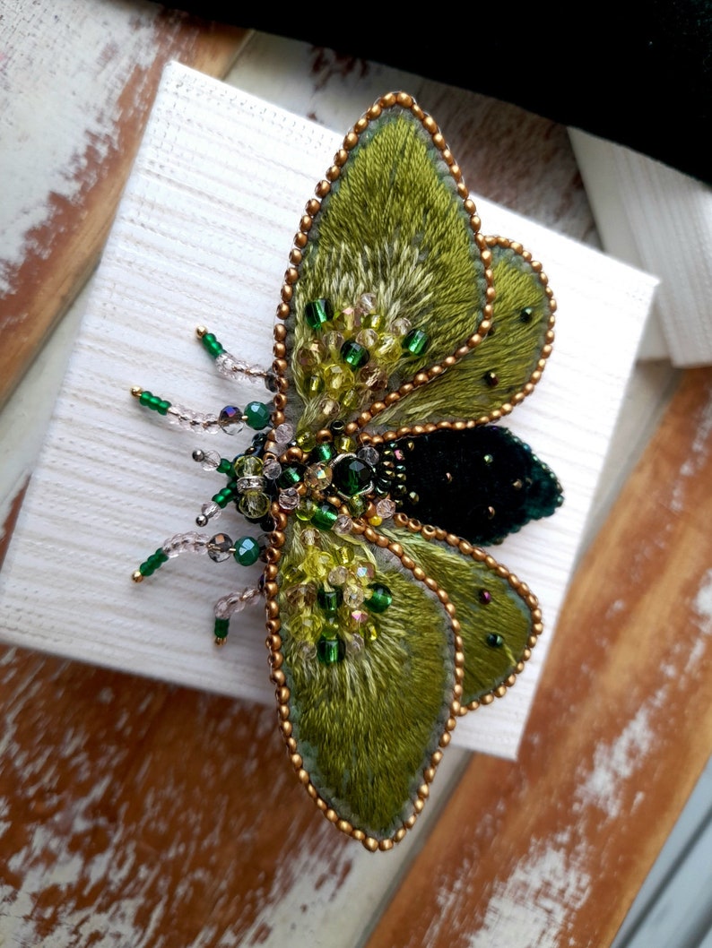 May include: A handcrafted, embroidered insect brooch. The wings are a textured green with gold beaded edges. The body is black velvet, and the insect features green and clear bead accents. The brooch is set on a white box.