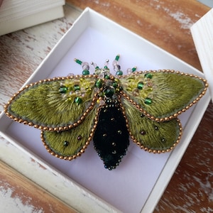 May include: A handcrafted moth brooch with olive green wings and a black body, embellished with beads and crystals. The wings have a textured, fur-like appearance, and the edges are outlined with gold-colored beads. The brooch is presented in a white box.