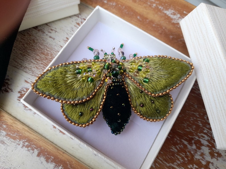 May include: A handcrafted moth brooch with olive green wings and a black body, embellished with beads and crystals. The wings have a textured, fur-like appearance, and the edges are outlined with gold-colored beads. The brooch is presented in a white box.