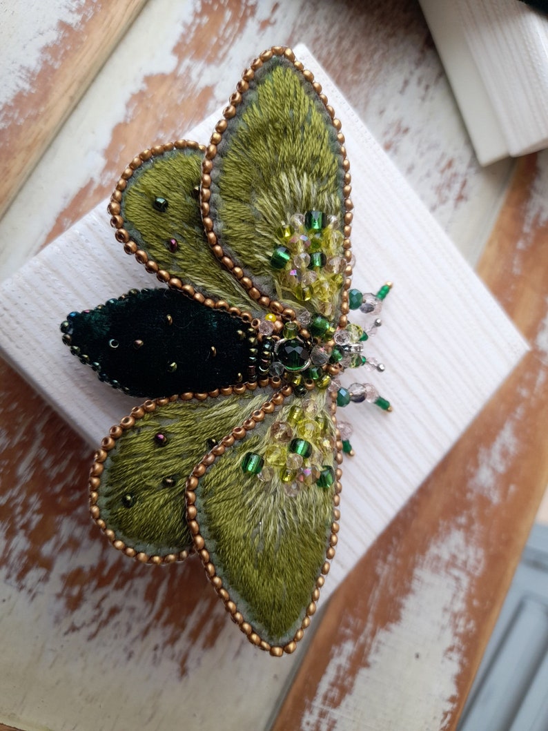 May include: An intricately embroidered moth brooch. The wings are a textured olive green, outlined with gold beads. The body is a deep forest green, with accents of green and clear beads. The brooch is set on a white surface.