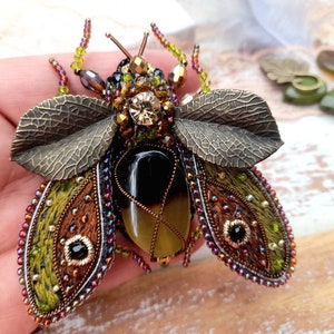 May include: A green and brown embroidered insect brooch with a black and yellow stone body and bronze leaf wings.