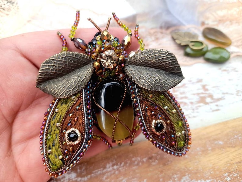 May include: A green and brown embroidered insect brooch with a black and yellow stone body and bronze leaf wings.