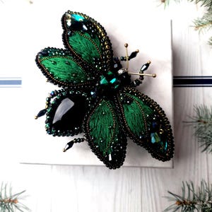 Pode incluir: Broche artesanal de inseto verde esmeralda com intrincado trabalho de contas e corpo de pedra preta. As asas são detalhadas com bordados verde escuro e contornadas com arame dourado. O broche está sobre uma superfície branca.