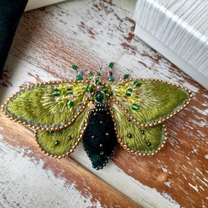 May include: A handcrafted, embroidered moth brooch. The wings are a textured, olive green with gold bead edging. The body is a deep, dark green velvet. Embellished with green and clear beads, this unique accessory adds a touch of nature-inspired elegance.
