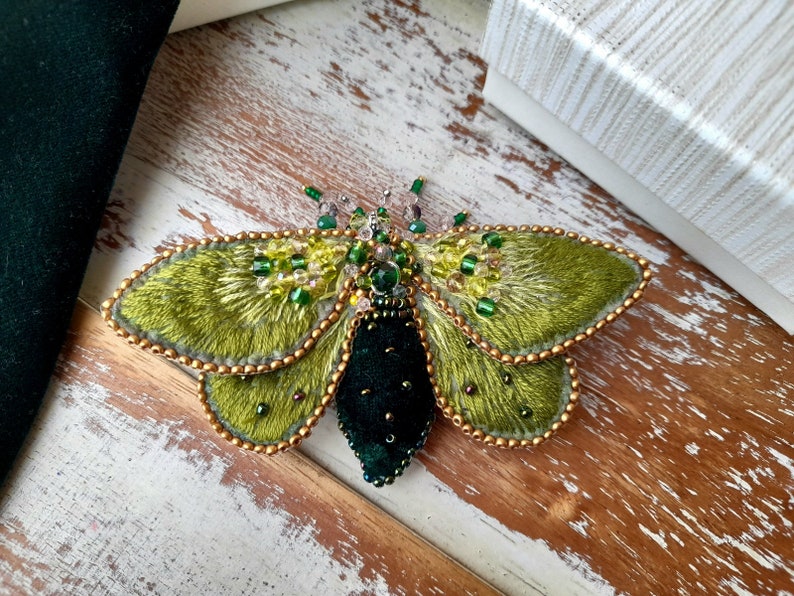May include: A handcrafted, embroidered moth brooch. The wings are a textured, olive green with gold bead edging. The body is a deep, dark green velvet. Embellished with green and clear beads, this unique accessory adds a touch of nature-inspired elegance.