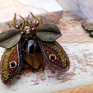 May include: A green, brown, and black embroidered insect brooch with a large, clear, faceted gemstone in the center. The wings are embellished with metallic leaves and beads.