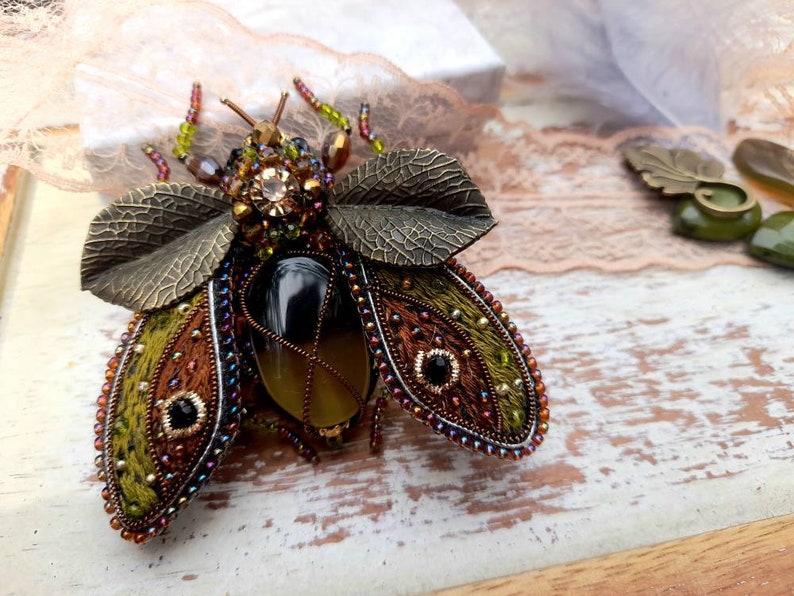 May include: A green, brown, and black embroidered insect brooch with a large, clear, faceted gemstone in the center. The wings are embellished with metallic leaves and beads.