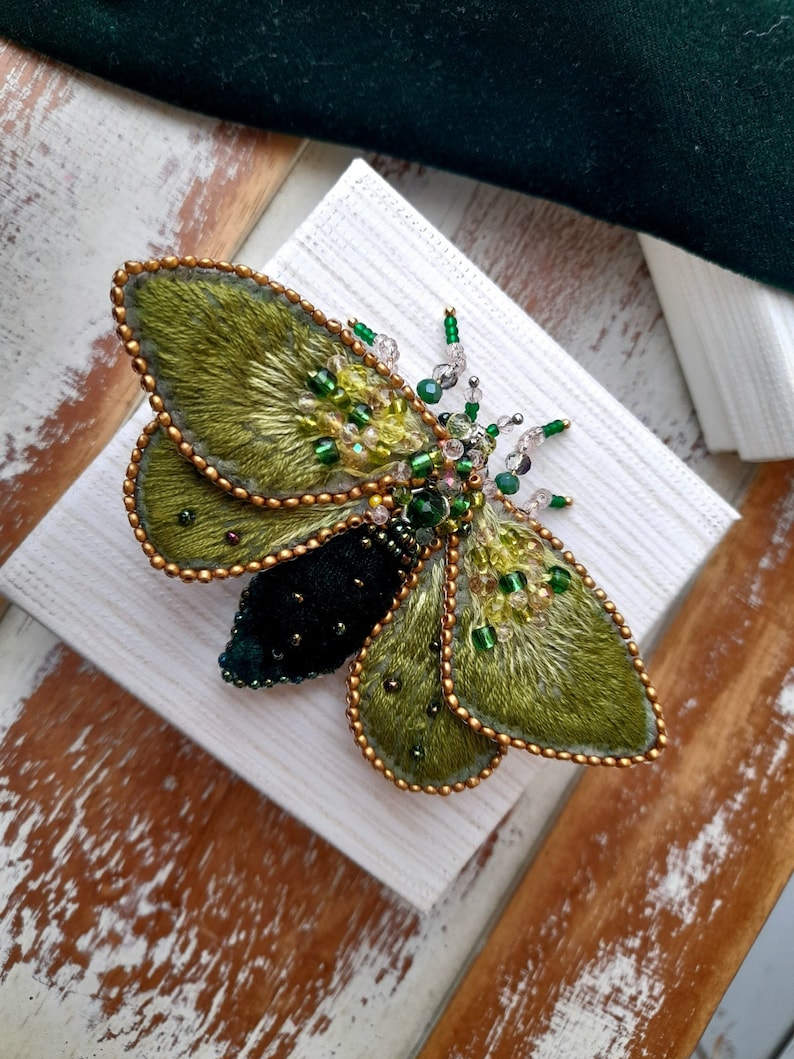 May include: A handcrafted, green and black insect brooch. The brooch features embroidered wings with gold beaded edges and is adorned with green and clear beads. The insect's body is a deep black velvet.