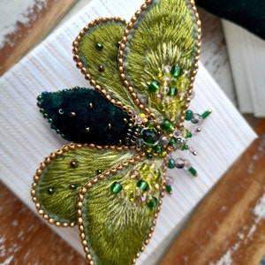 May include: A handcrafted, embroidered butterfly brooch. The wings are a vibrant green with gold beaded edges. The body is a dark green velvet, and the brooch is adorned with green and clear beads. A unique, artistic accessory.