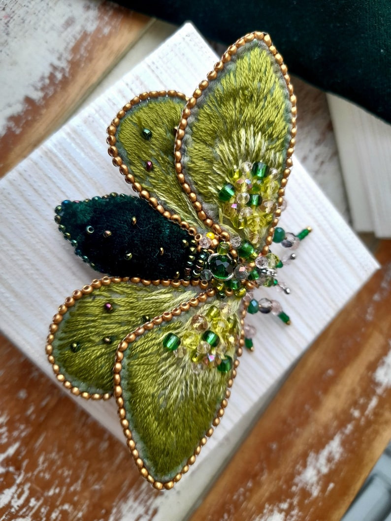 May include: A handcrafted, embroidered butterfly brooch. The wings are a vibrant green with gold beaded edges. The body is a dark green velvet, and the brooch is adorned with green and clear beads. A unique, artistic accessory.