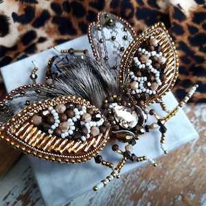May include: A handcrafted insect brooch featuring intricate beadwork in shades of gold, brown, and white. The wings are made of sheer lace with metallic edges, while the body includes a central teardrop crystal, dark fur, and beaded legs.