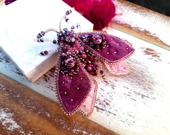 May include: A purple and pink velvet moth brooch with beaded wings and rhinestones. The brooch is handmade and features intricate details.