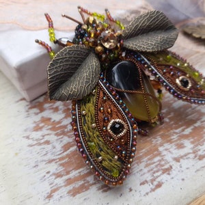 May include: A beaded and embroidered brooch in the shape of a beetle with green, brown, and gold accents. The beetle has a large, clear, yellow stone in its center.