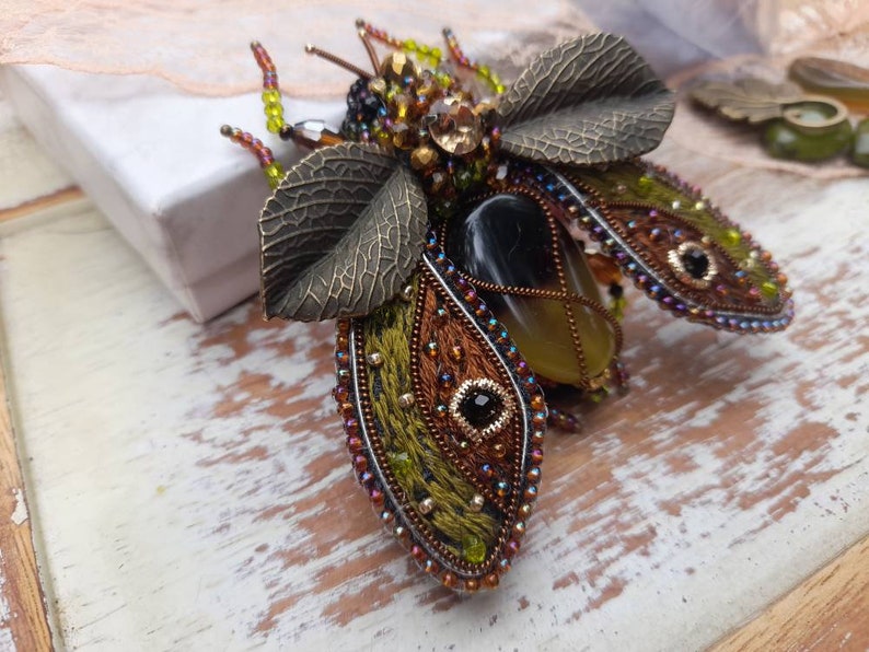 May include: A beaded and embroidered brooch in the shape of a beetle with green, brown, and gold accents. The beetle has a large, clear, yellow stone in its center.