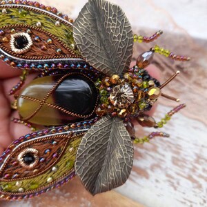 May include: A detailed, beaded and embroidered brooch in the shape of a beetle. The beetle has iridescent wings, a black and brown body, and a large, clear crystal on its back.