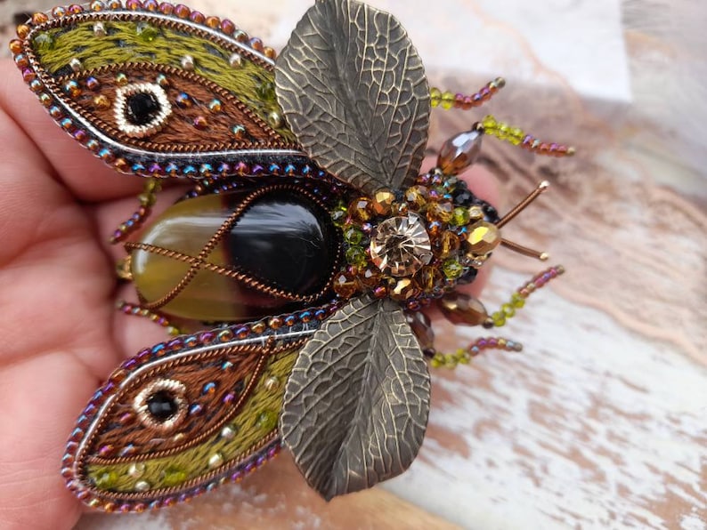 May include: A detailed, beaded and embroidered brooch in the shape of a beetle. The beetle has iridescent wings, a black and brown body, and a large, clear crystal on its back.